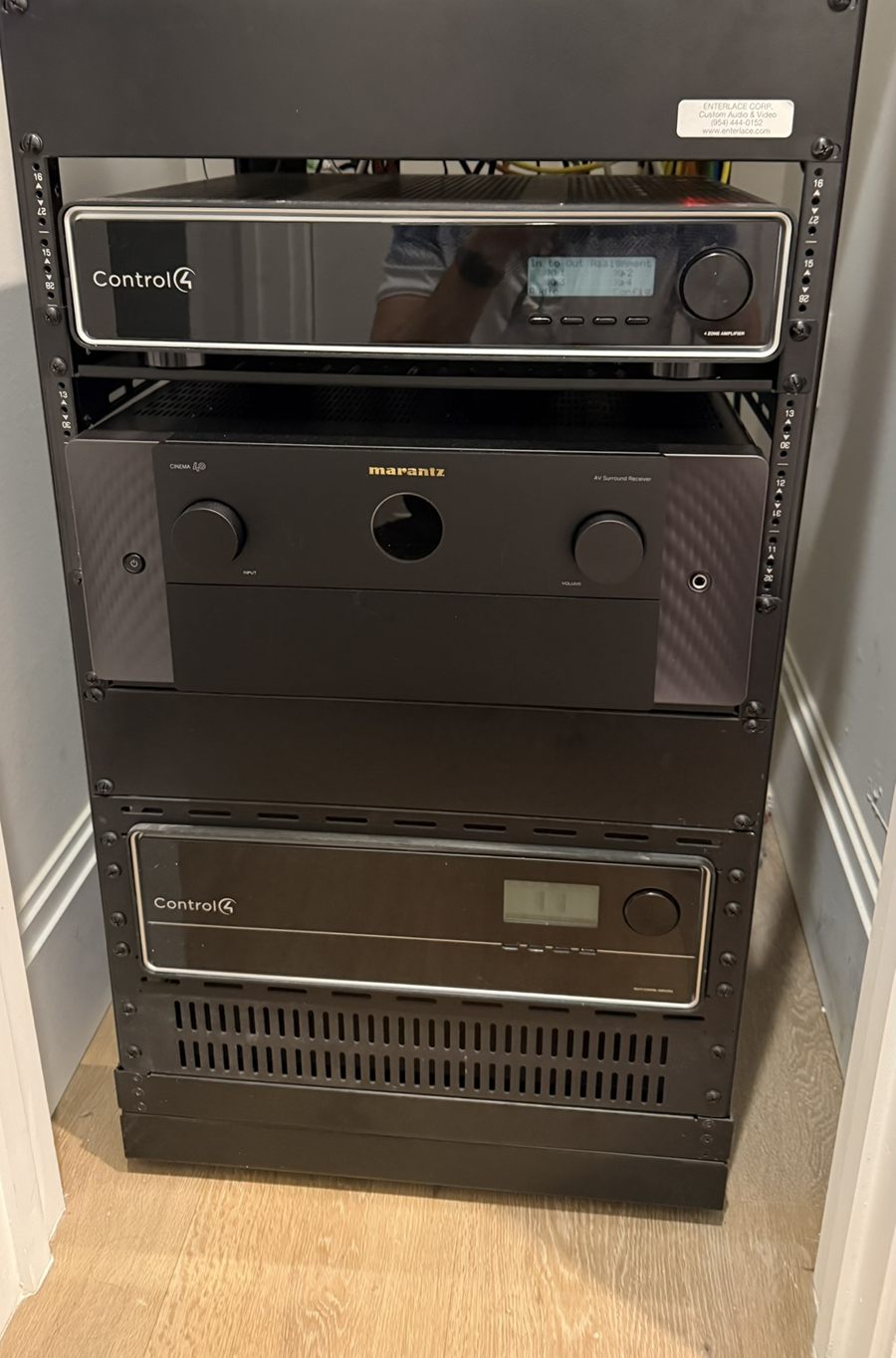 A black equipment rack standing in a closet, holding a Control4 four-zone amplifier, a Marantz CINEMA 40 AV surround receiver and a second Control4 amplifier, with an Enterlace Corp Custom Audio and Video sticker on the blank panel above them