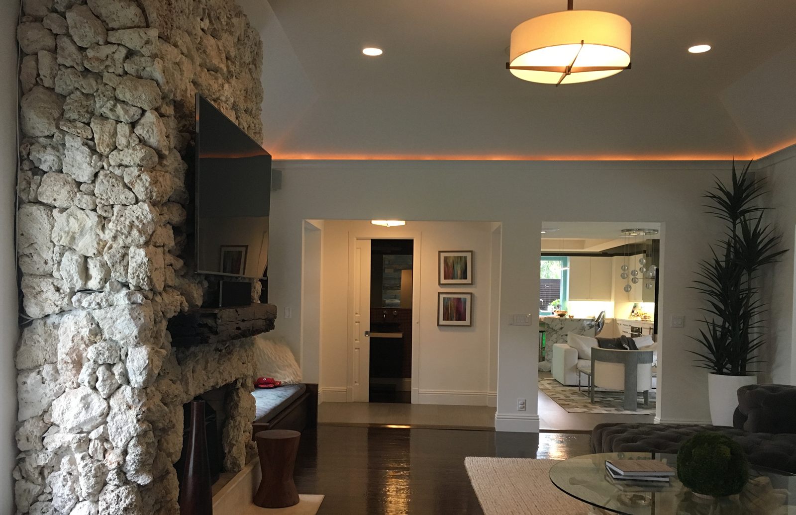 An open-plan living room lit by warm cove lighting running around the edge of a tray ceiling, with recessed downlights, a lit drum pendant, a coral rock chimney breast with a wall-mounted television, dark hardwood floors, and a kitchen visible through an opening beyond