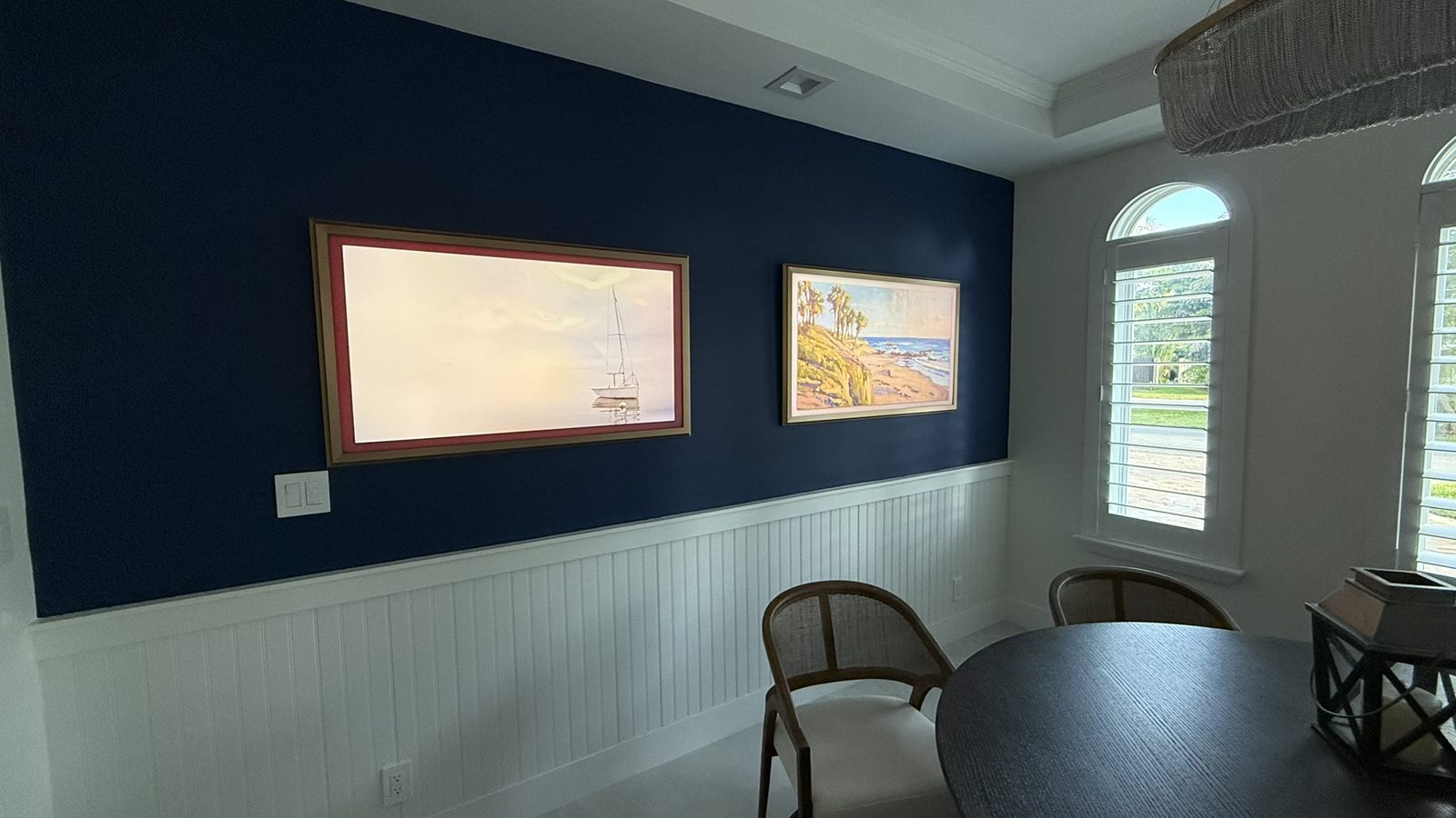 A large wall-mounted television in art mode displaying a painting of a sailboat, set in a gold and red picture frame on a navy dining room wall above white beadboard wainscoting, with a smaller framed coastal painting hung beside it