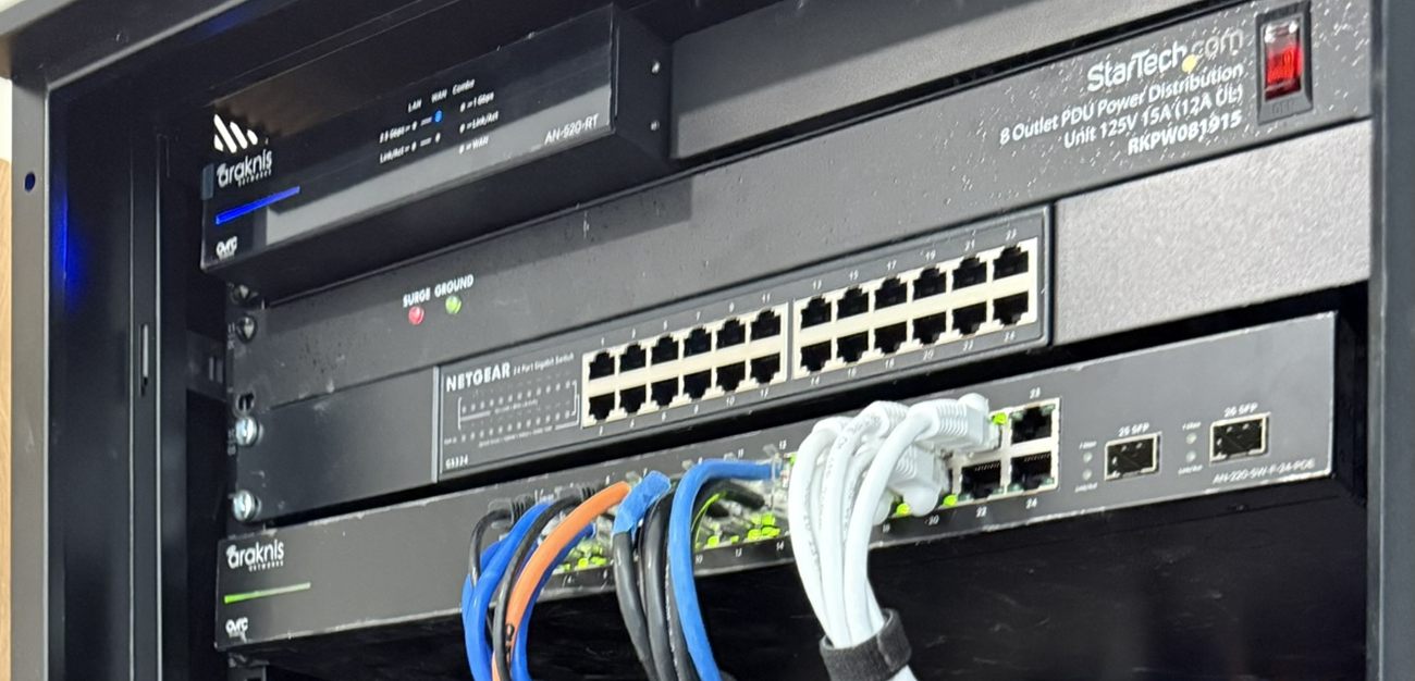 The upper section of a wall-mounted network cabinet holding an Araknis router, a StarTech eight-outlet rack power distribution unit, a NETGEAR 24-port gigabit switch and an Araknis 24-port PoE switch, with blue, orange, black and white patch cords gathered into a velcro strap